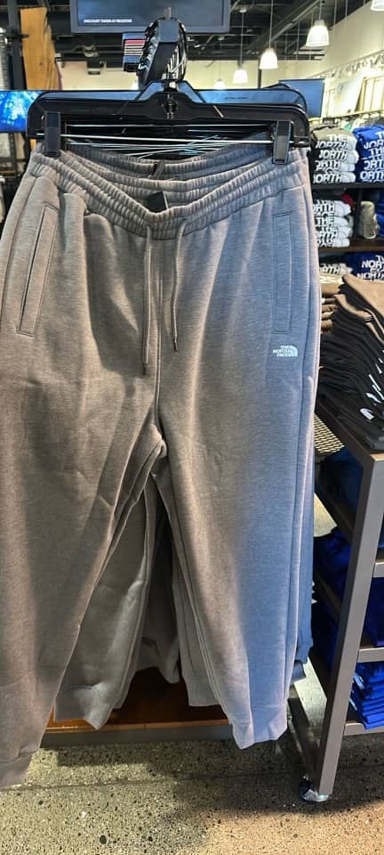 Calças jogger de fleece The North Face e, ao lado, mesas com moletons e camisetas com logo.